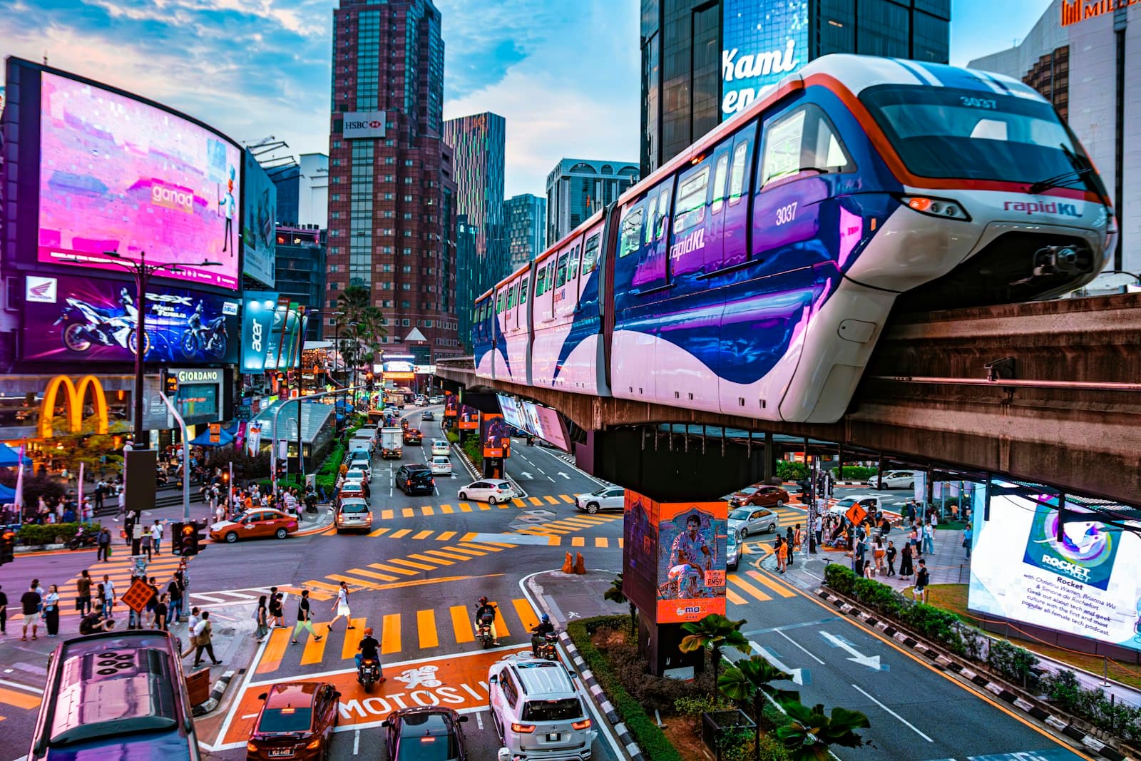 A Rapid KL monorail train curving above a busy Bukit Bintang junction at dusk, with pedestrians on the striped crossing and lit billboards on the towers behind